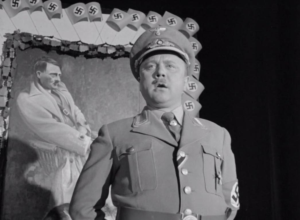 Mann in Uniform steht vor Hitler-Gemälde, umgeben von Hakenkreuzfahnen.