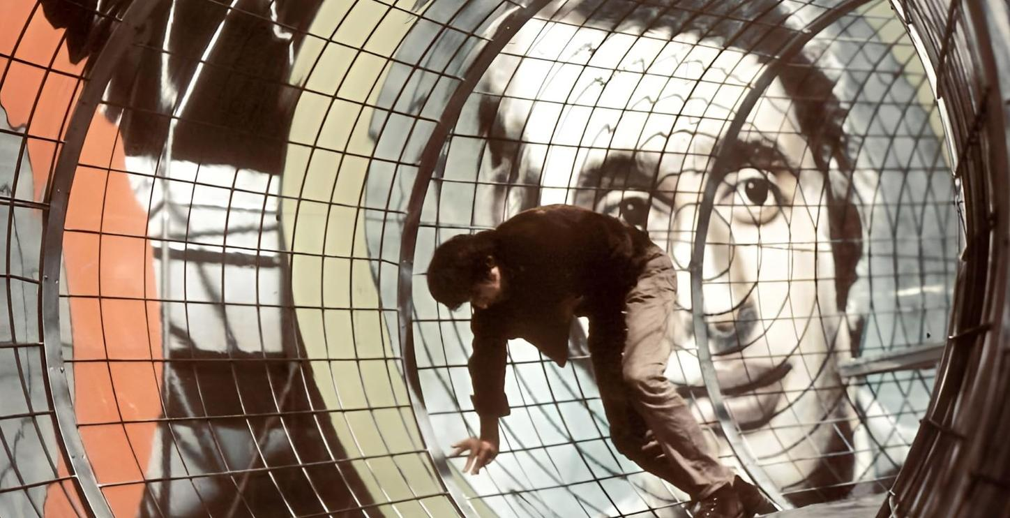 Mann in dunkler Kleidung klettert in einem Gitterrad vor einem riesigen Gesichtsbild.