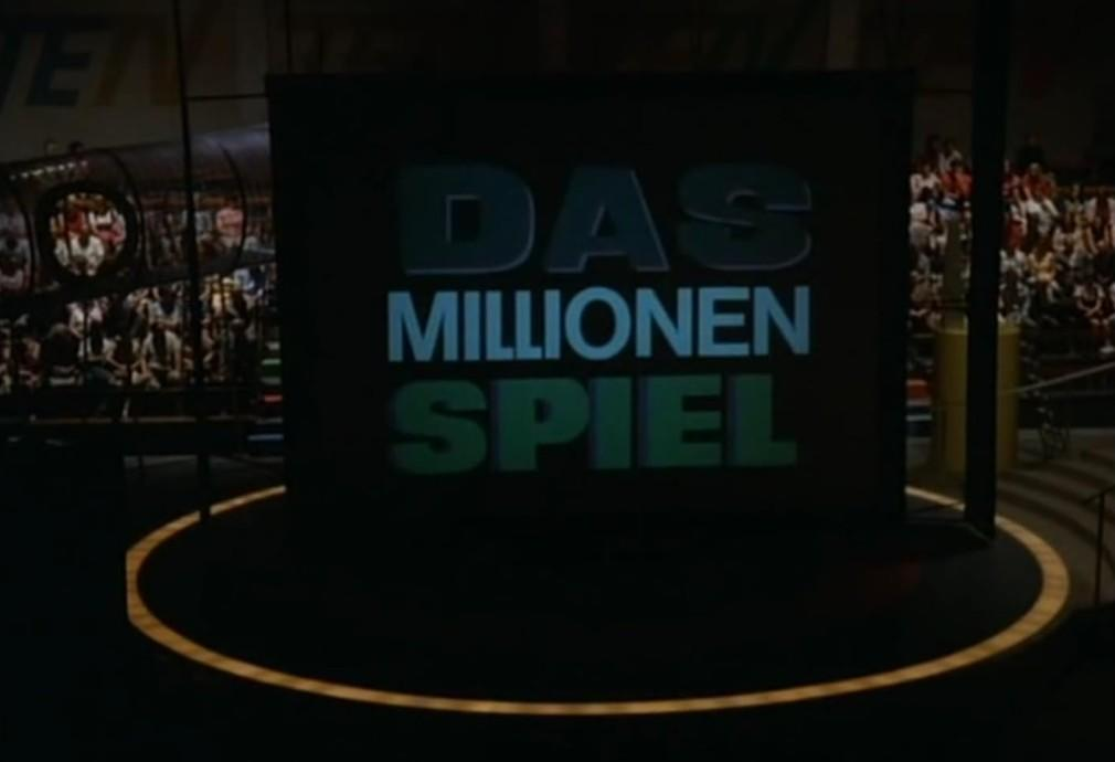 Die Aufschrift "DAS MILLIONEN SPIEL" auf einer dunklen Bühne mit Publikum im Hintergrund.