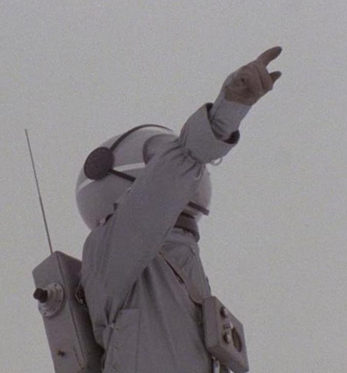 Ein Astronaut in einem grauen Raumanzug zeigt nach oben, mit einem Rucksack und Antenne.