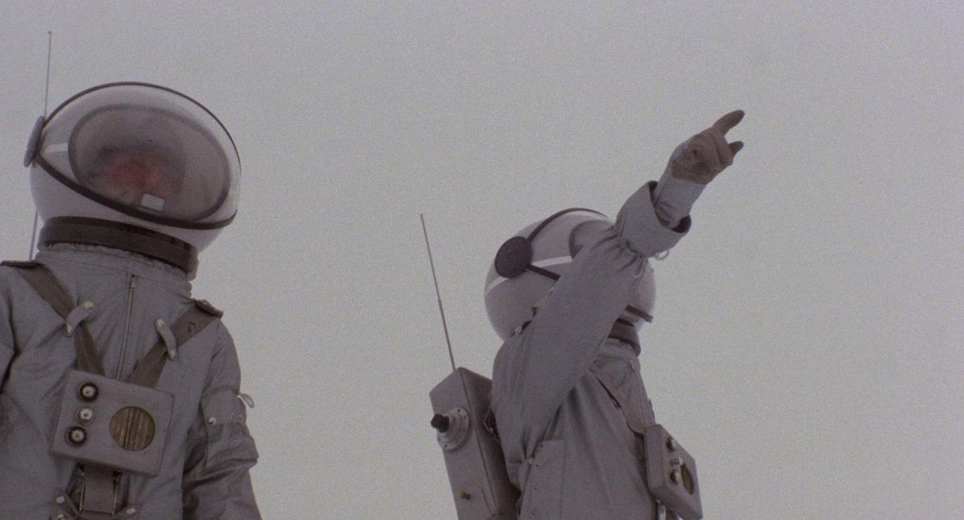 Zwei Astronauten in grauen Anzügen mit klaren Helmen. Einer zeigt nach oben, der andere schaut voraus.