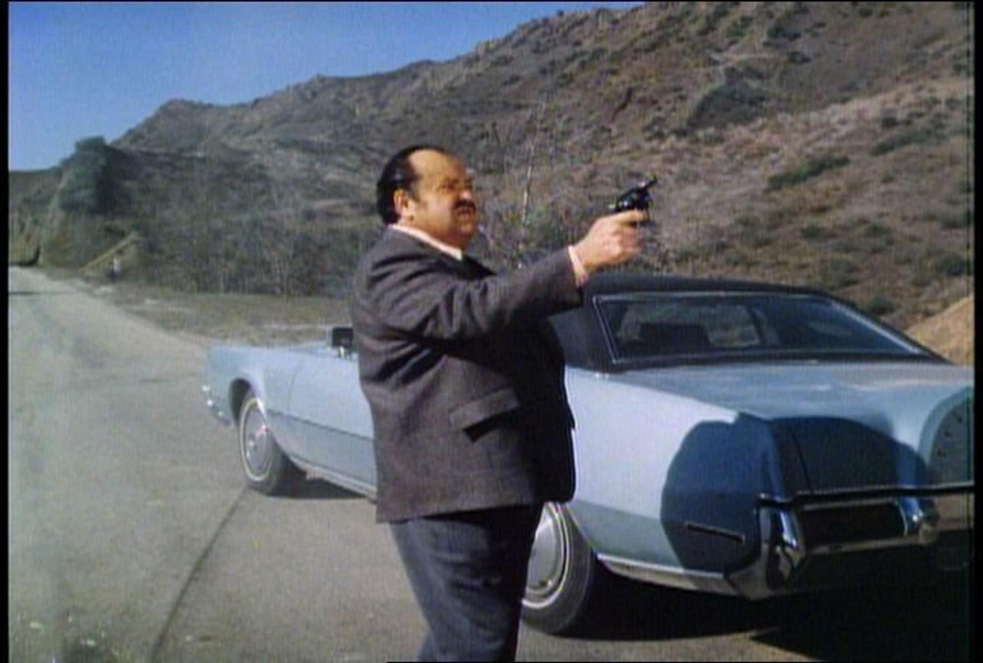 Ein Mann in einem Anzug zielt mit einer Pistole neben einem Auto in einer bergigen Landschaft.