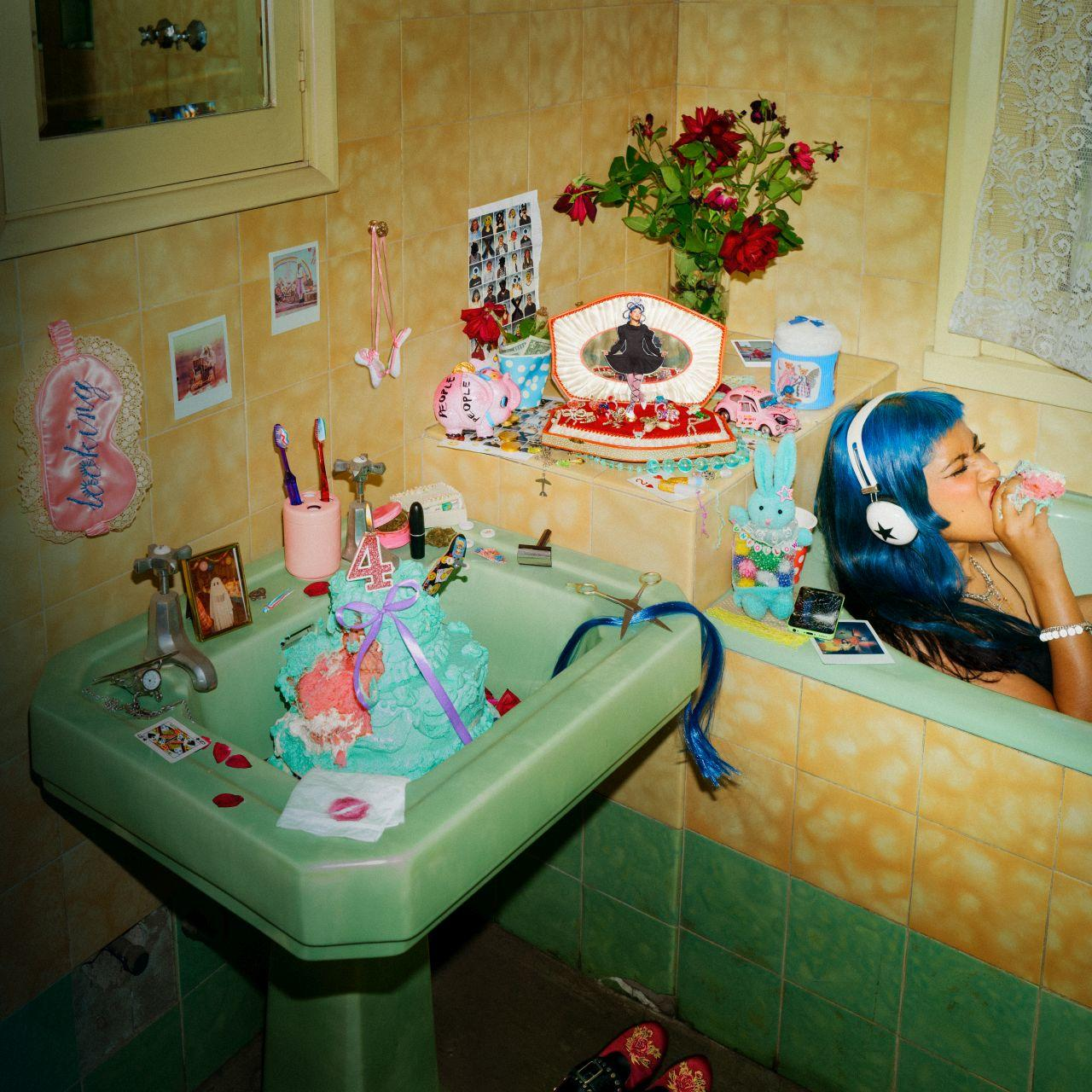 Eine Frau mit blauen Haaren sitzt in einer Badewanne und hört Musik. Ein Waschbecken ist mit Kuchen und Dekorationen überfüllt.