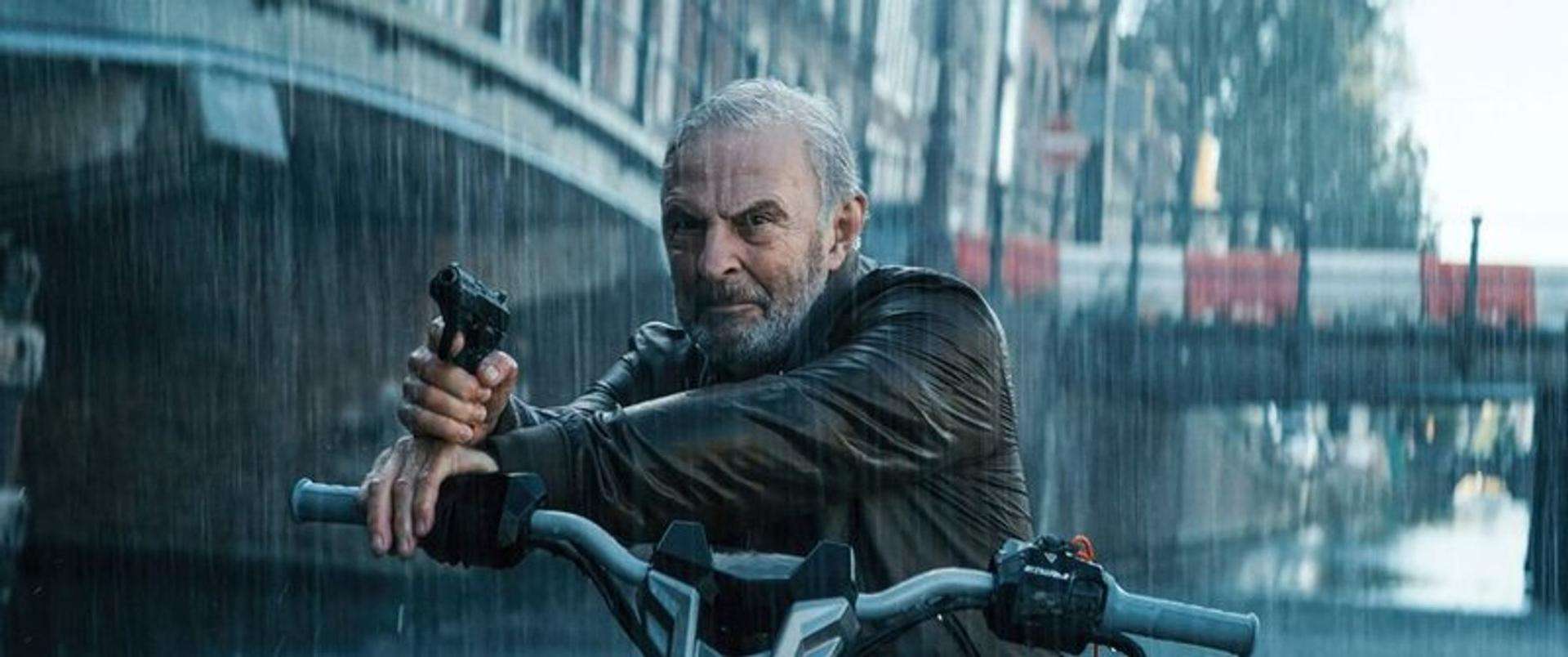 Ein Mann mit grauem Haar auf einem Motorrad hält eine Pistole im Regen.