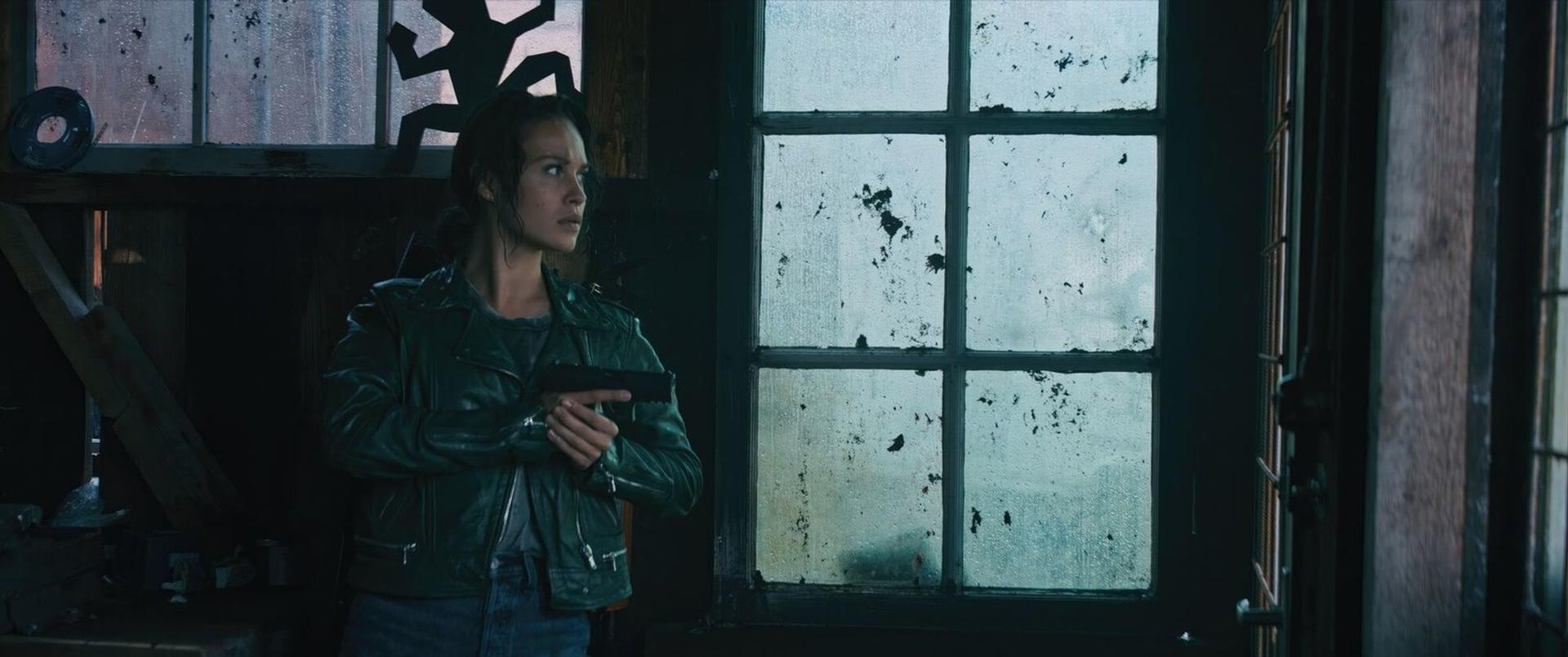 Eine Frau in Lederjacke hält eine Pistole in einem dunklen Raum mit einem schmutzigen Fenster.