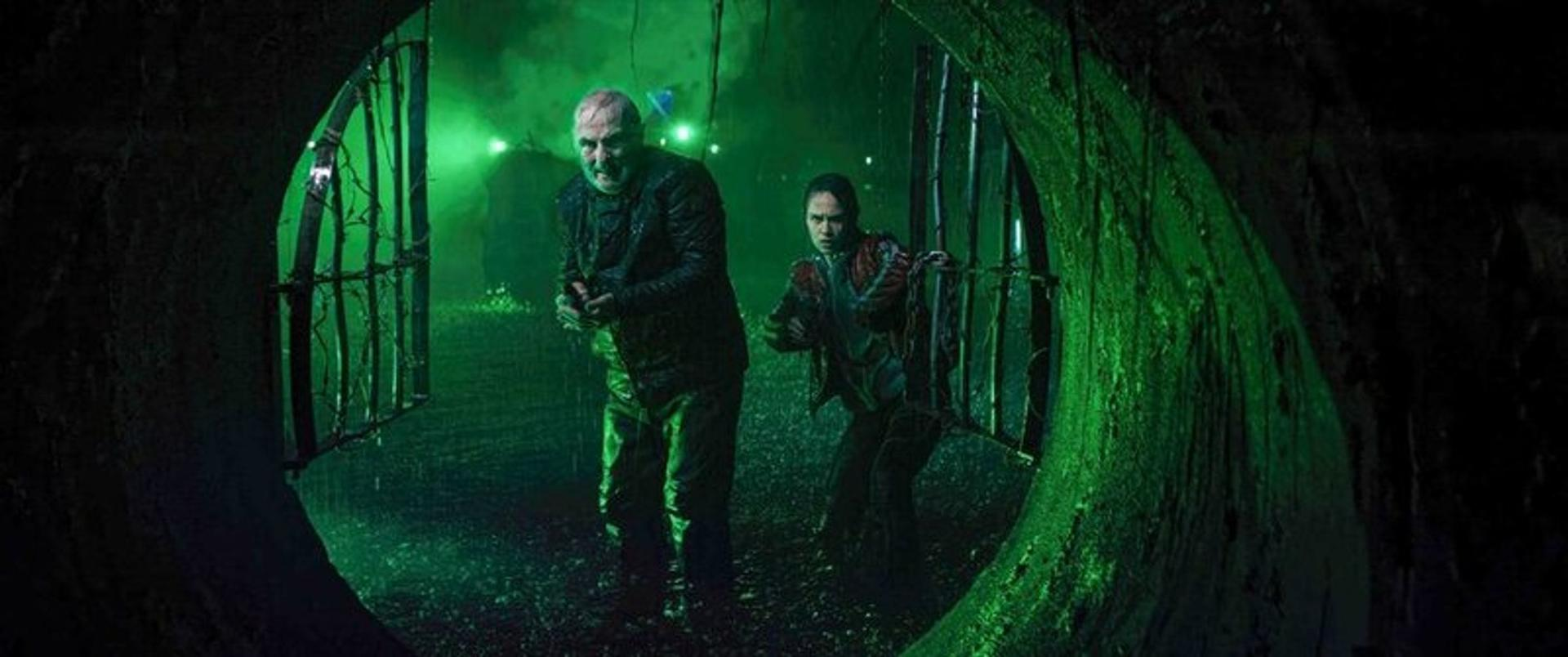 Zwei Personen stehen in grünem Licht hinter einem Gitter im Regen.