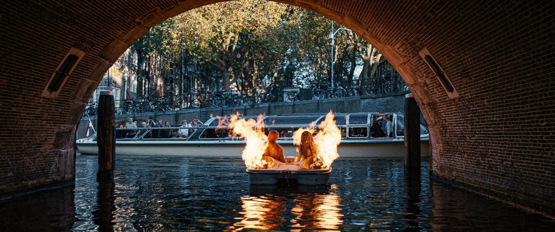 Zwei Personen in einem Boot mit Flammen, die von einem Flussufer unter einer Ziegelbrücke fahren.