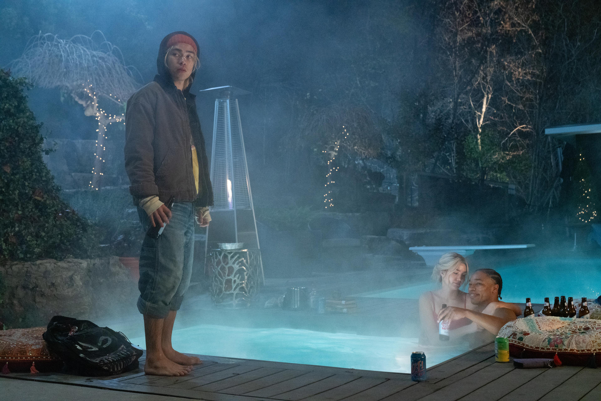Ein junger Mann steht an einem Pool, während sich ein Paar nachts im Wasser umarmt.