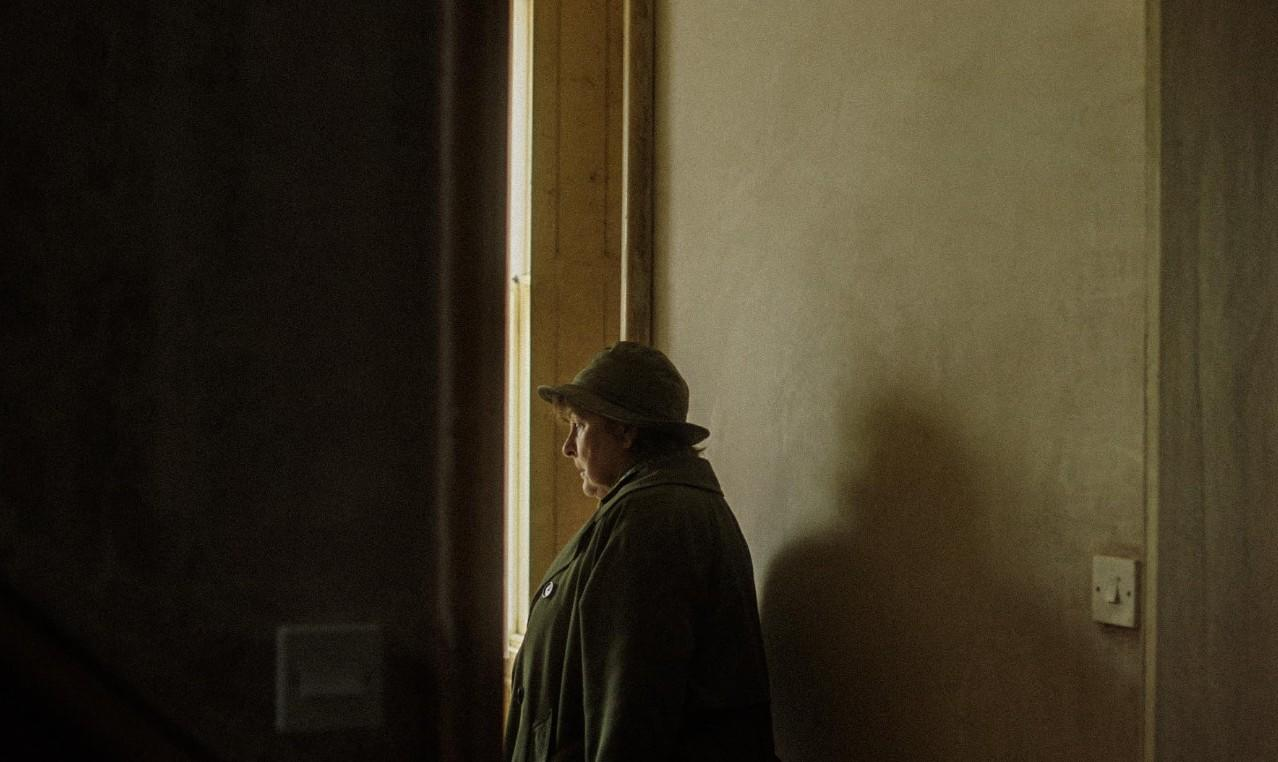 Eine Person im Hut steht vor einem Fenster. Schatten an der Wand.