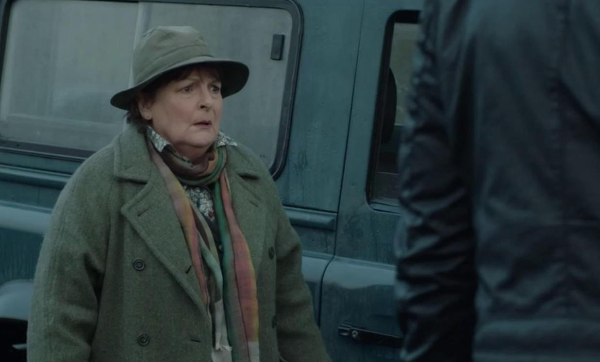 Eine Frau mit Hut und Mantel steht neben einem dunklen Fahrzeug.