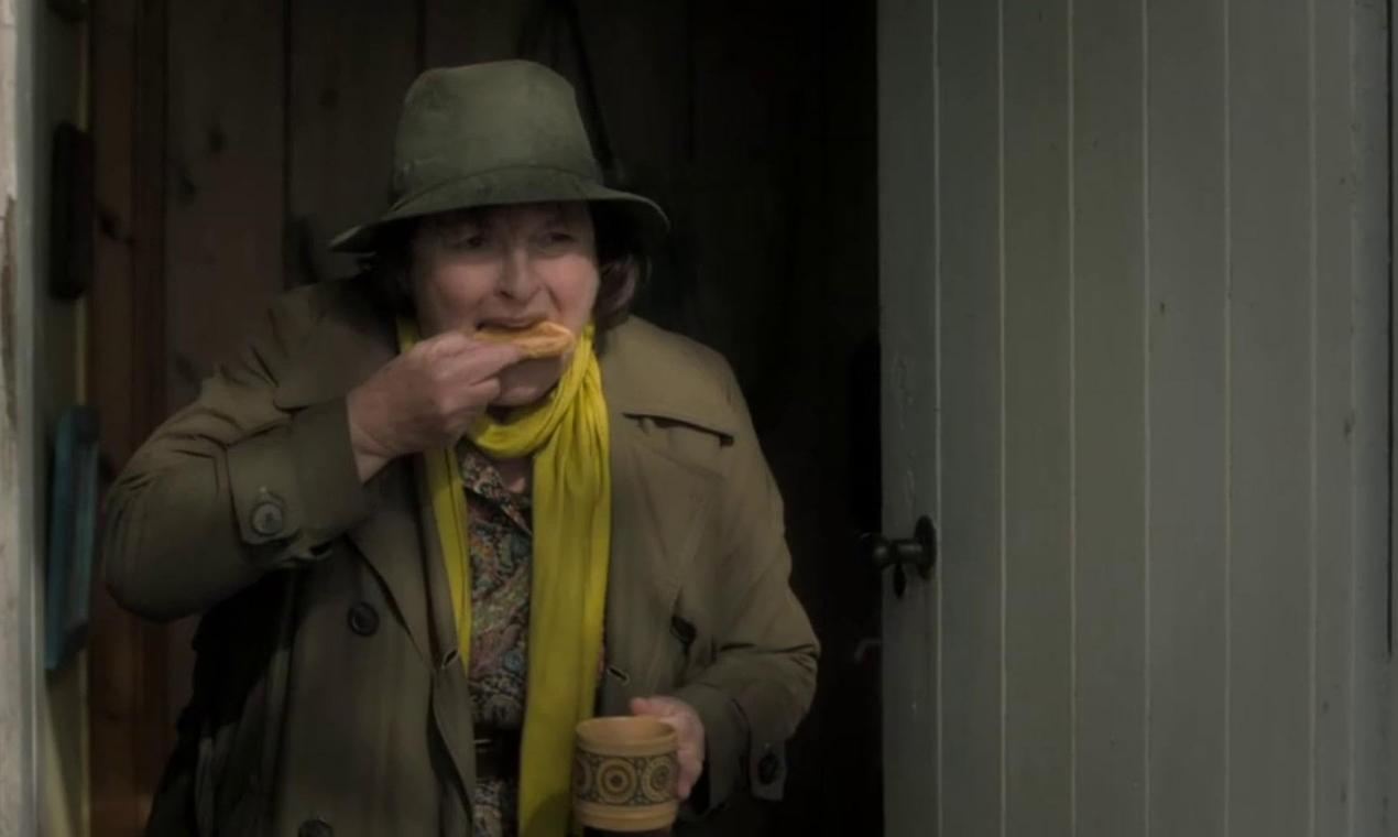 Eine Frau mit Hut und Schal isst ein Stück Essen und hält einen Becher.