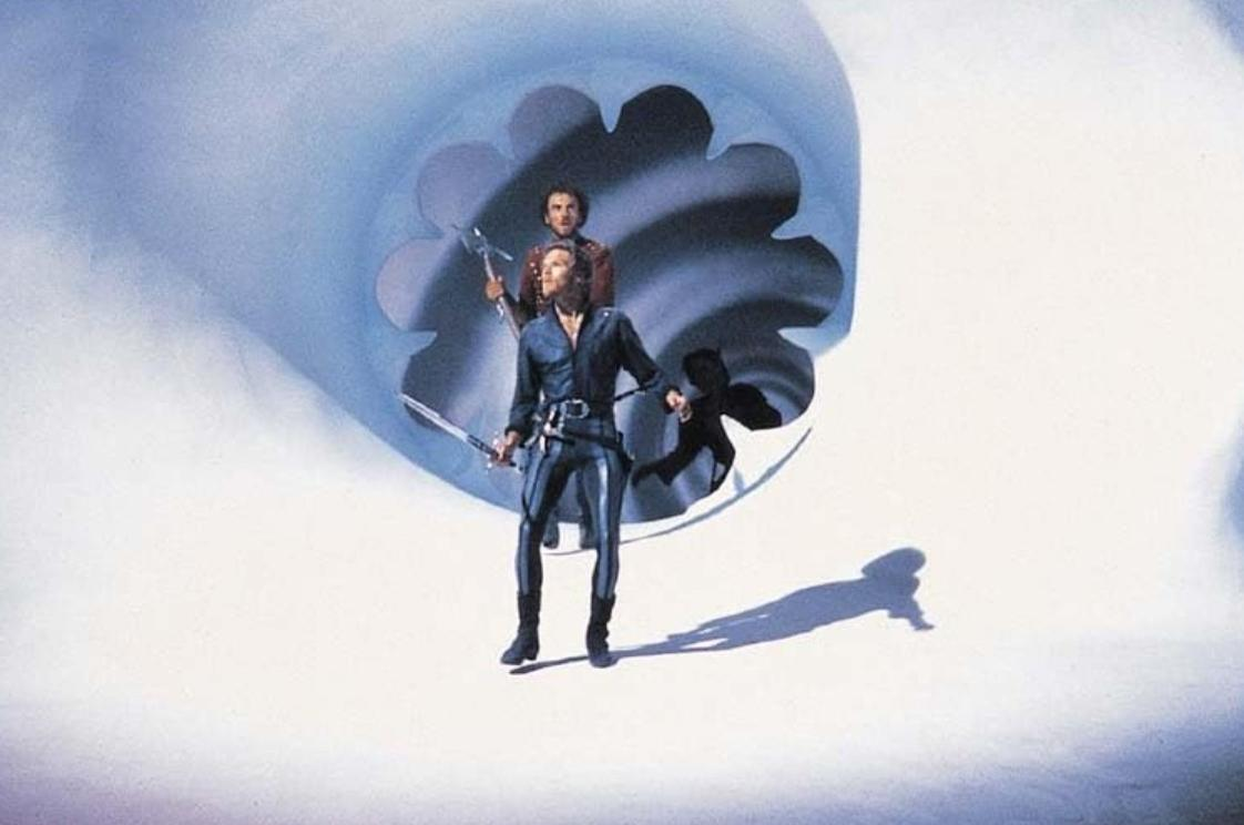 Zwei Männer in mittelalterlicher Kleidung stehen in einem futuristischen, blau beleuchteten Tunnel und halten Schwerter.