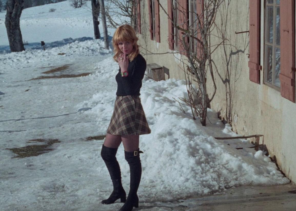 Eine Frau mit blonden Haaren steht im Schnee vor einem Haus mit roten Fensterläden.