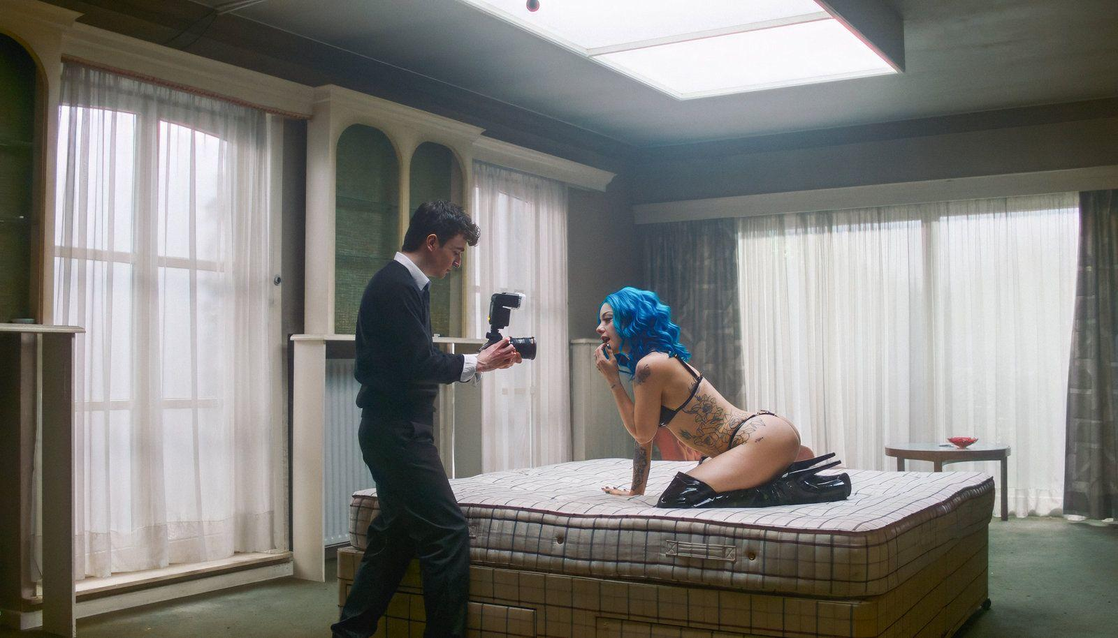 Ein Mann mit Kamera fotografiert eine Frau mit blauen Haaren auf einem Bett.
