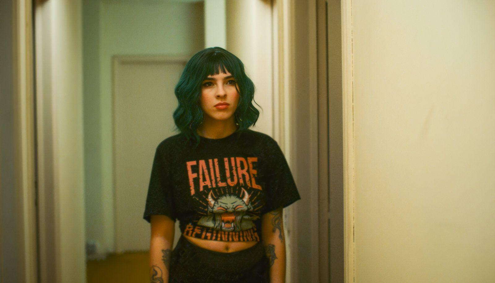 Eine junge Frau mit tealgrünen Haaren trägt ein schwarzes T-Shirt mit dem Aufdruck FAILURE.