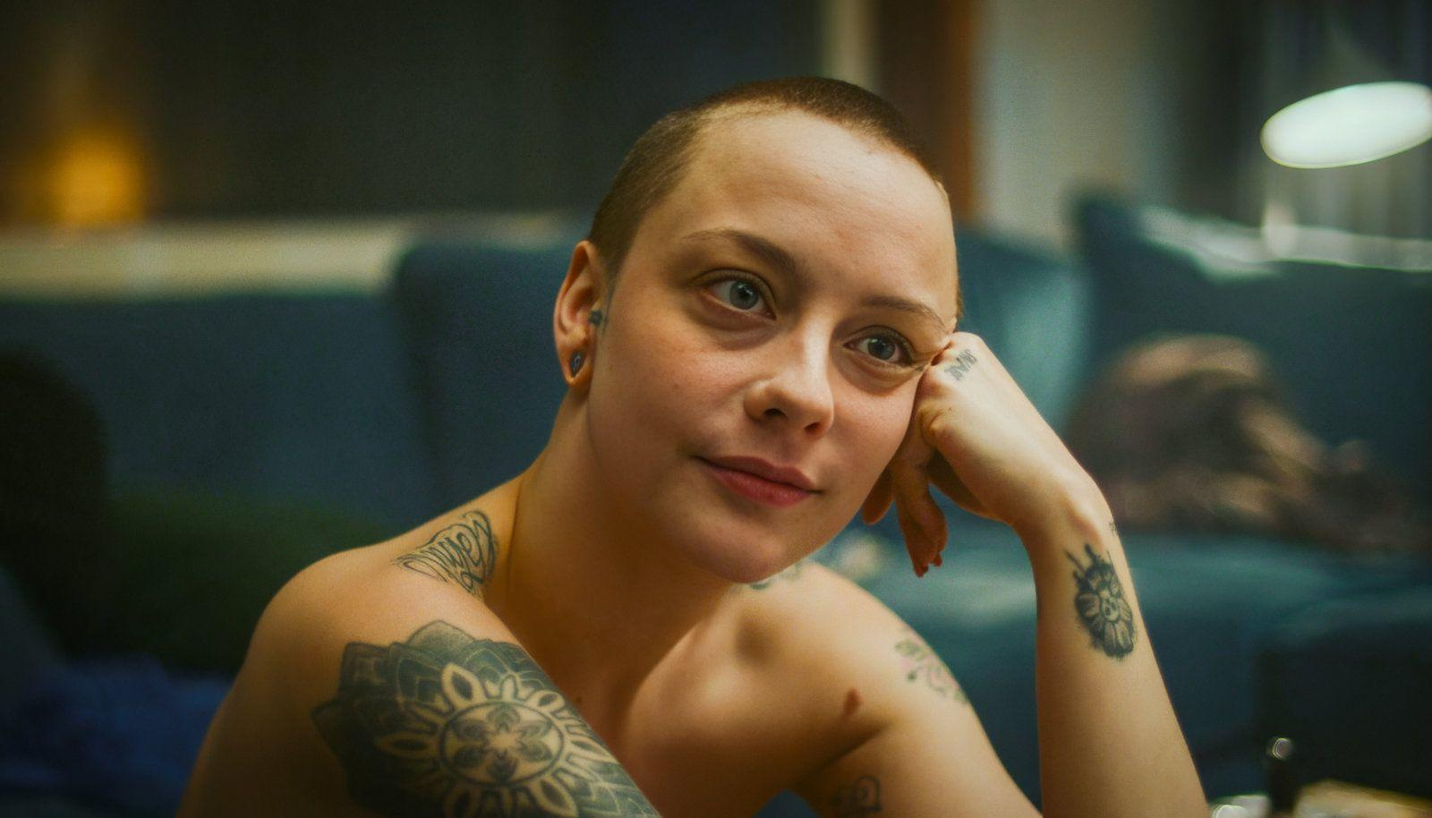 Eine Frau mit kurzgeschorenen Haaren und Tattoos stützt ihr Kinn auf ihre Hand.