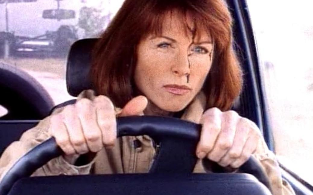 Eine Frau mit blutigem Gesicht fährt ein Auto. Sie hat rote Haare.