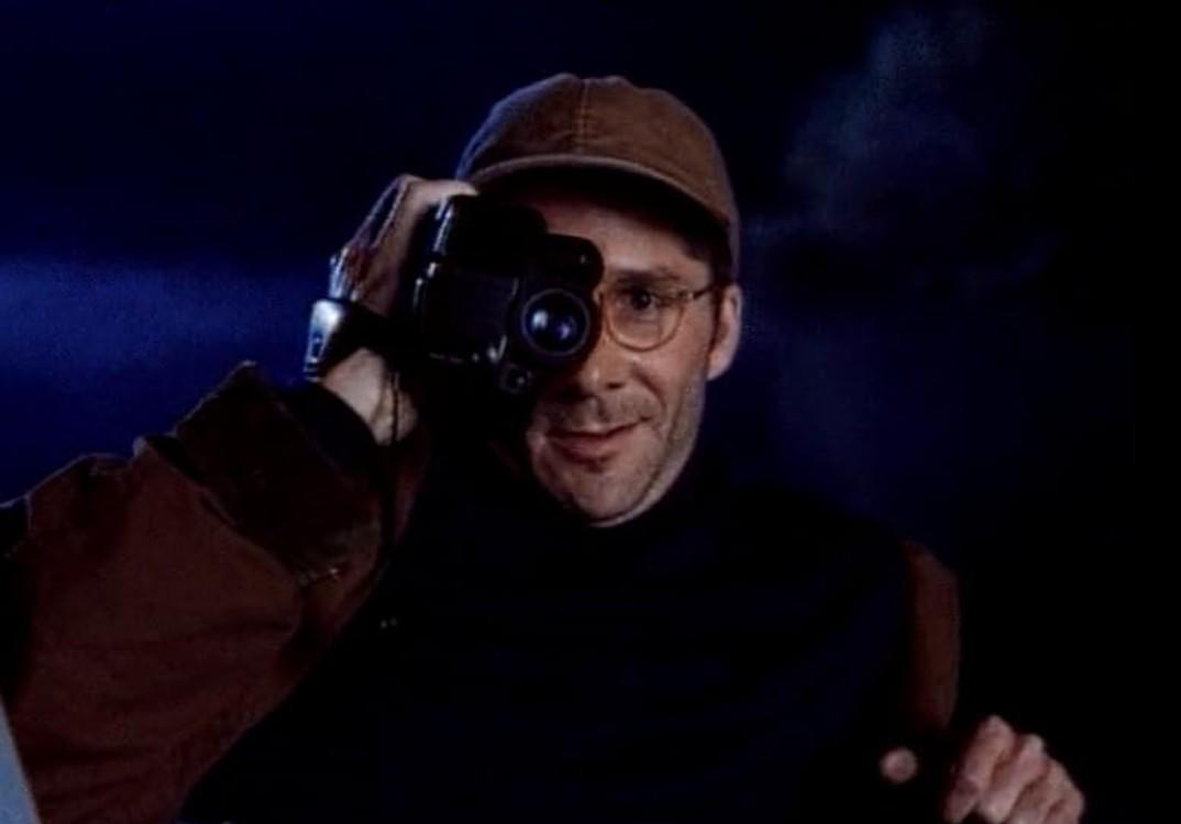 Ein Mann mit Brille und Kappe filmt mit einer Kamera, dunkler Hintergrund.