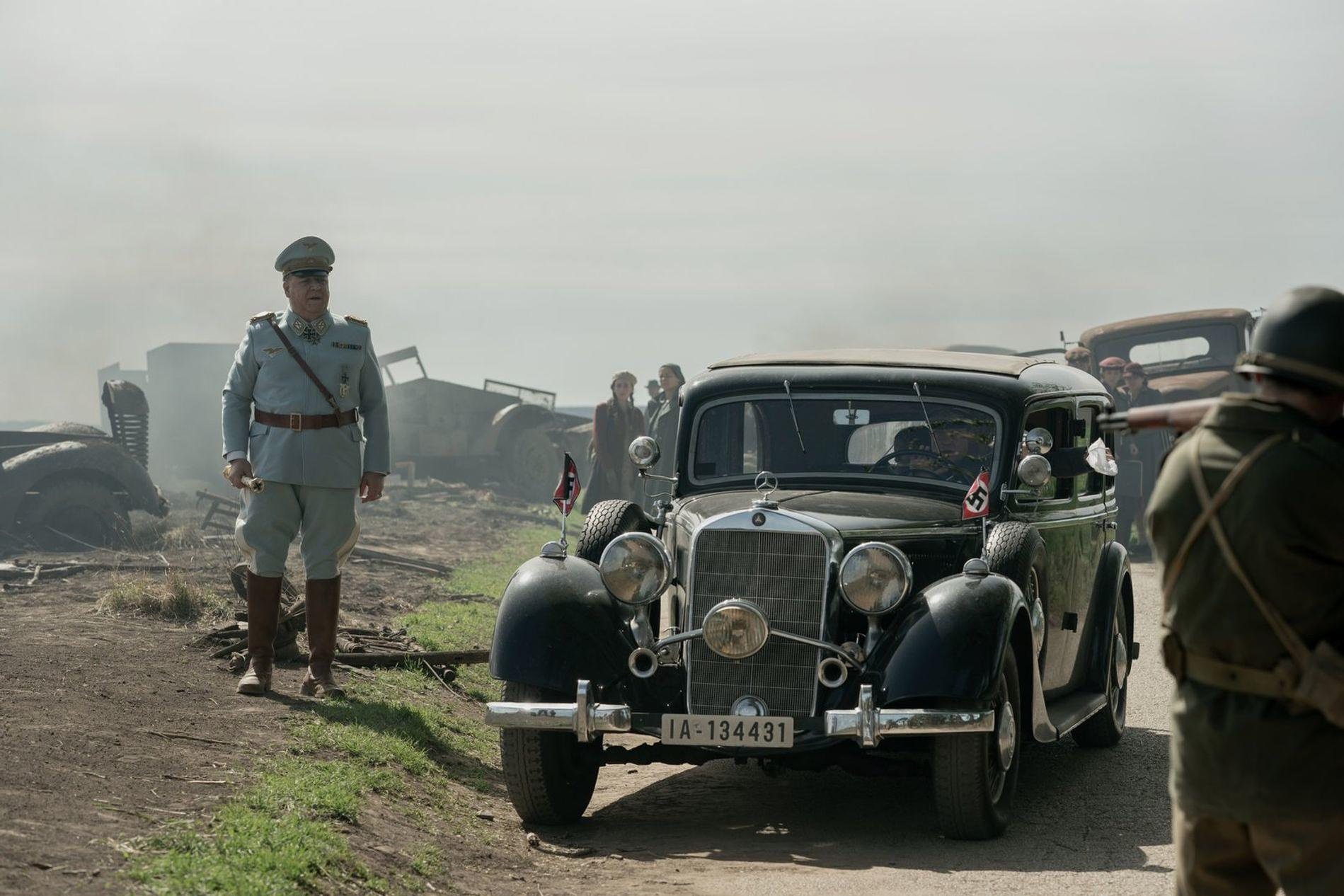 Ein Mann in Militäruniform steht neben einem Auto. Im Hintergrund sind weitere Fahrzeuge und Personen zu sehen.