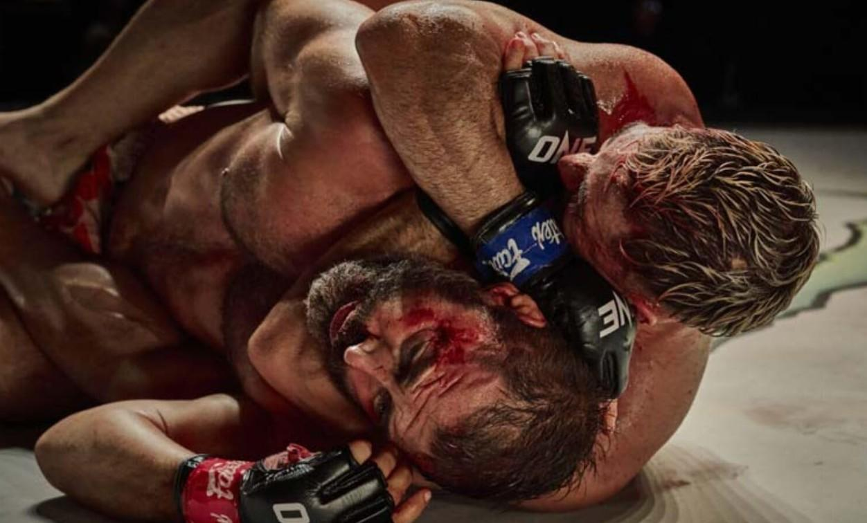 Zwei blutende MMA-Kämpfer im Clinch auf dem Boden, einer hält den anderen fest.