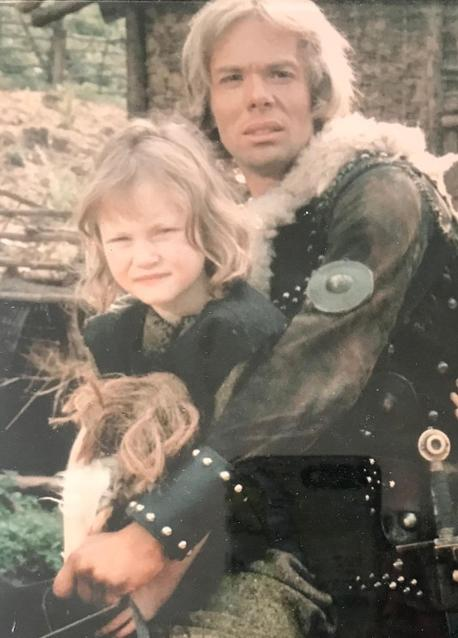 Ein Mann mit blonden Haaren umarmt ein kleines Mädchen vor einer Holzwand.