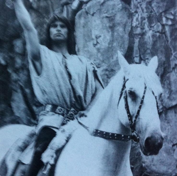 Mann mit erhobener Hand reitet auf einem weißen Pferd vor Felsen.