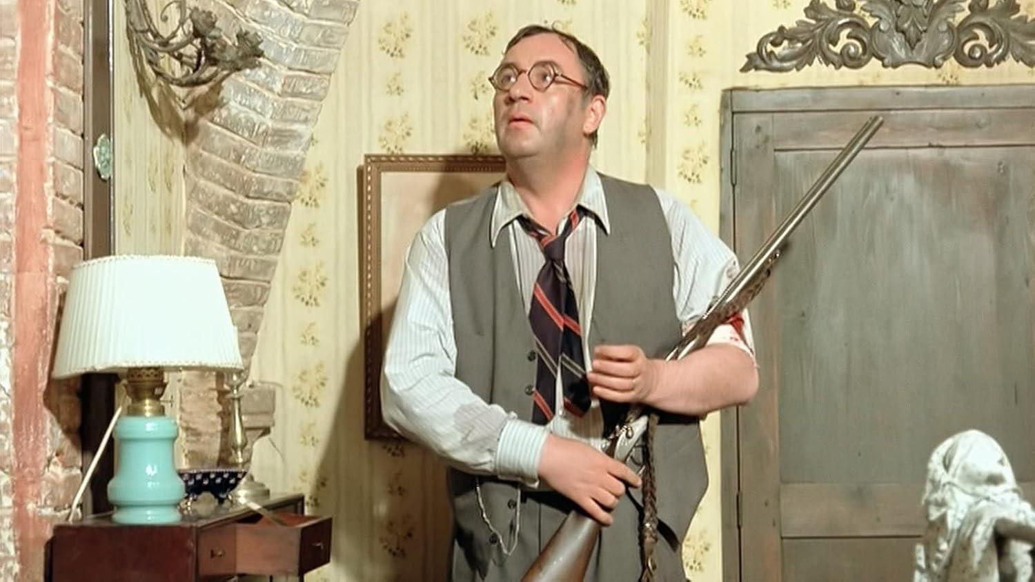 Ein Mann mit Brille hält eine Schrotflinte in einem Raum mit gemusterten Tapeten.