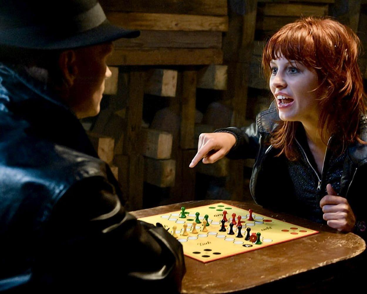 Eine Frau mit roten Haaren zeigt auf ein Brettspiel, das von zwei Personen gespielt wird.