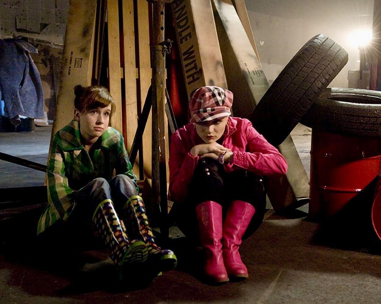 Zwei junge Frauen sitzen auf dem Boden, eine in Grün, die andere in Pink, umgeben von Holzkisten und Reifen.