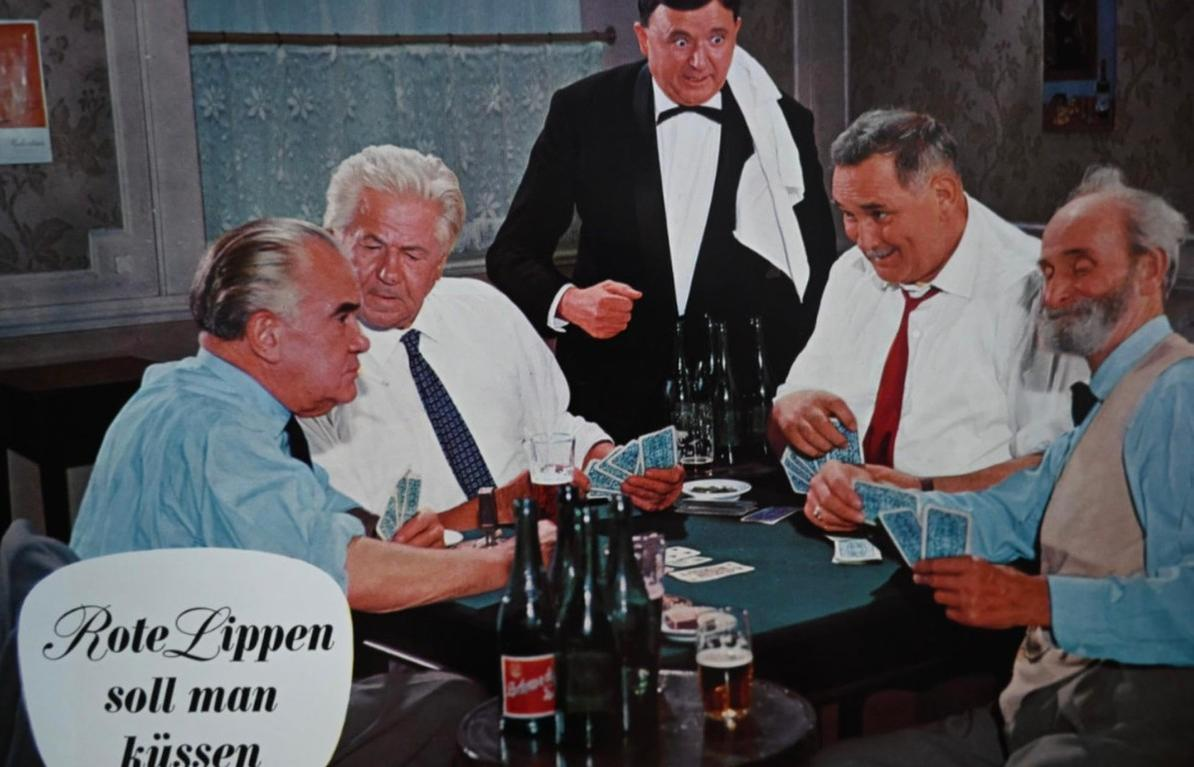 Fünf Männer spielen Karten an einem Tisch mit Bierflaschen und Gläsern.