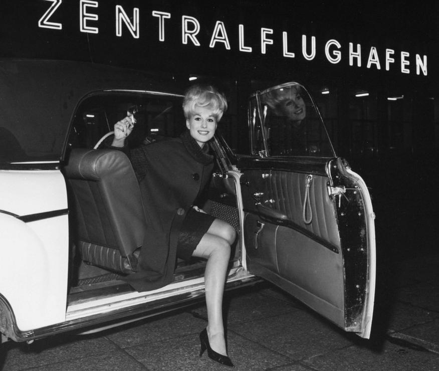 Frau in dunklem Kleid und Umhang steigt nachts aus Oldtimer. Neonschild ZENTRALFLUGHAFEN.
