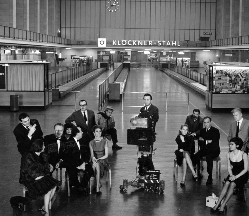 Gruppe von Menschen posiert vor einer Filmkamera in einer Halle mit "KLÖCKNER-STAHL"-Schild.