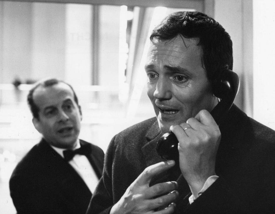 Mann im Anzug spricht am Telefon, ein anderer Mann im Smoking hinter ihm.