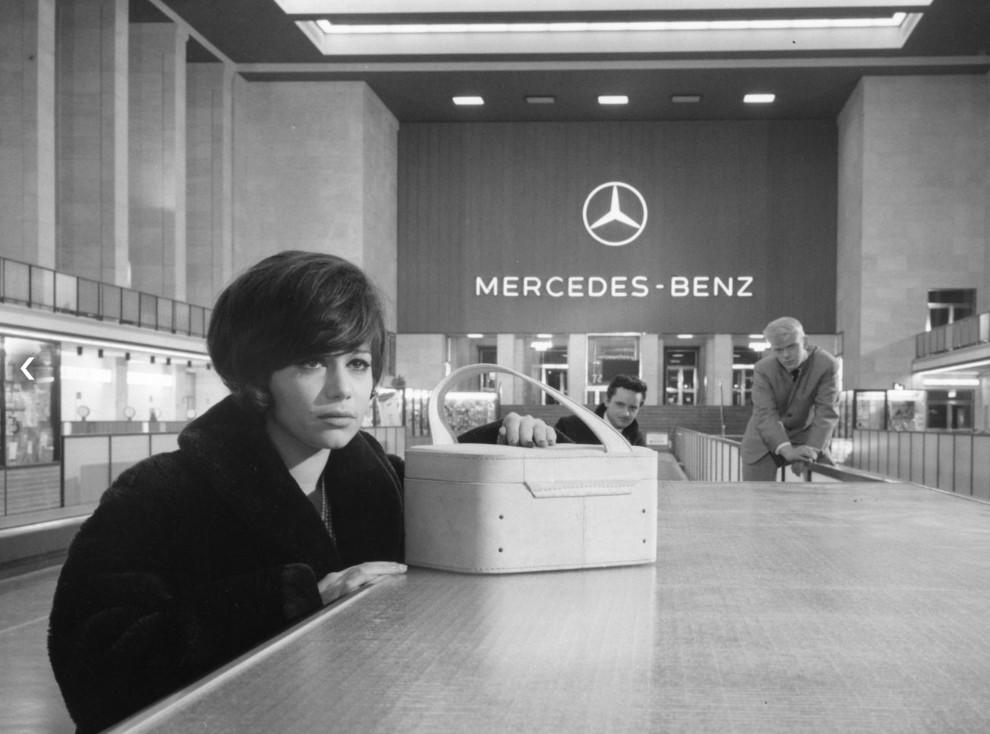 Frau mit Tasche vor Mercedes-Benz Logo. Zwei Männer im Hintergrund.