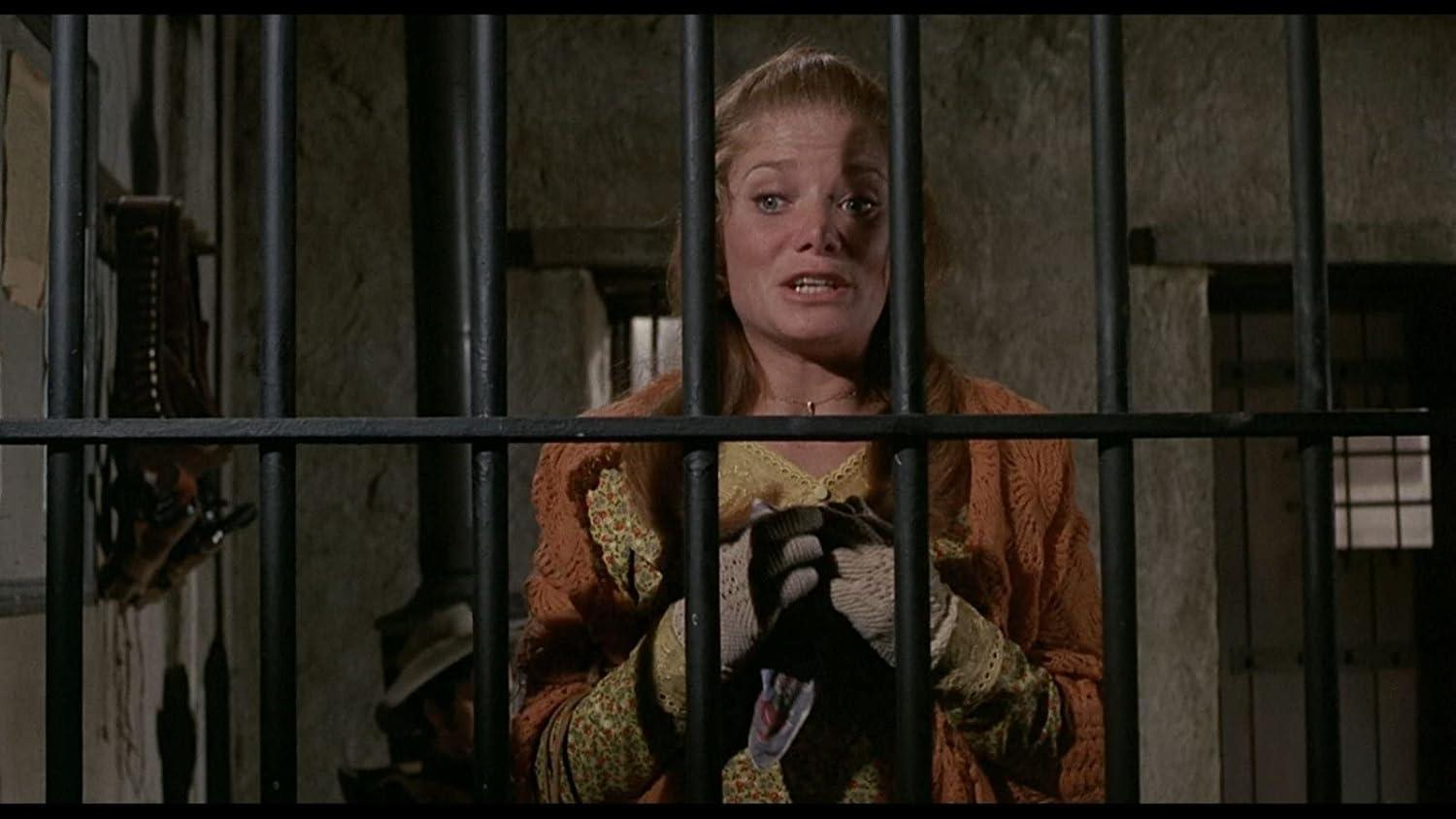 Eine Frau mit blonden Haaren hinter Gittern, sie trägt ein orangefarbenes Strickoberteil.