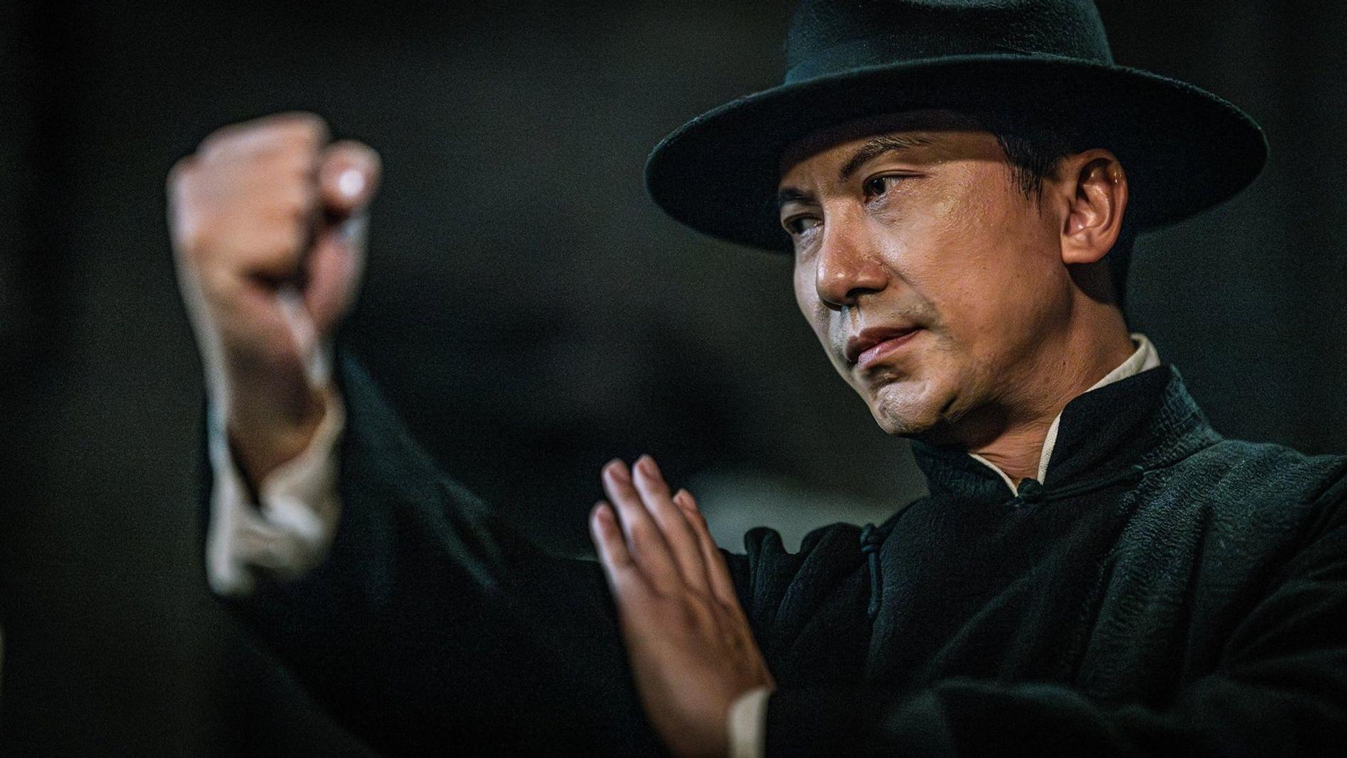 Ein Mann in schwarzem Hut und traditioneller chinesischer Kleidung zeigt eine Kampfkunstpose.