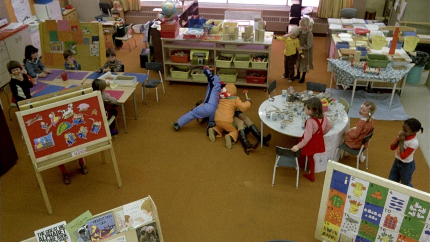 Kinder spielen in einem Klassenzimmer mit Tischen, Stühlen und einem Spielbereich.
