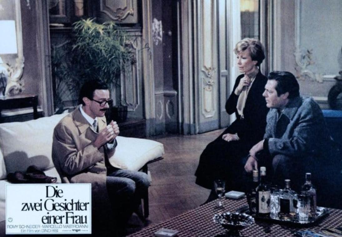 Drei Personen sitzen in einem opulenten Raum. Ein Mann links, eine Frau in der Mitte und ein Mann rechts.