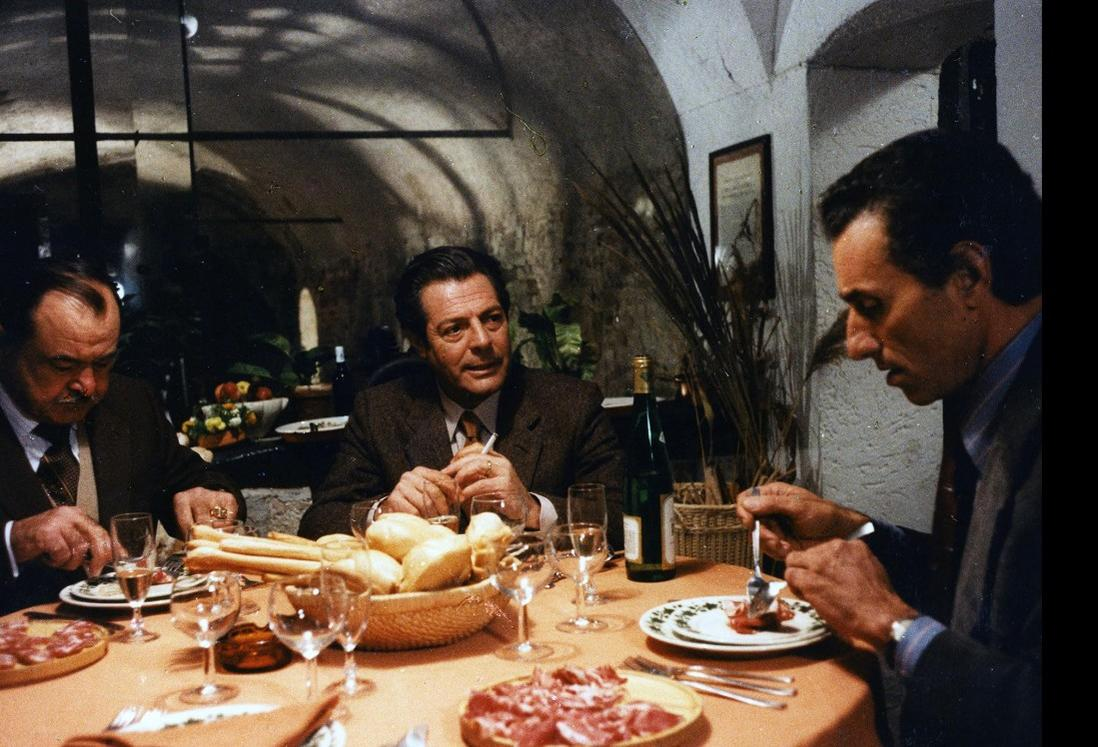 Drei Männer sitzen an einem Tisch mit Brot, Wurst und Wein. Einer raucht.