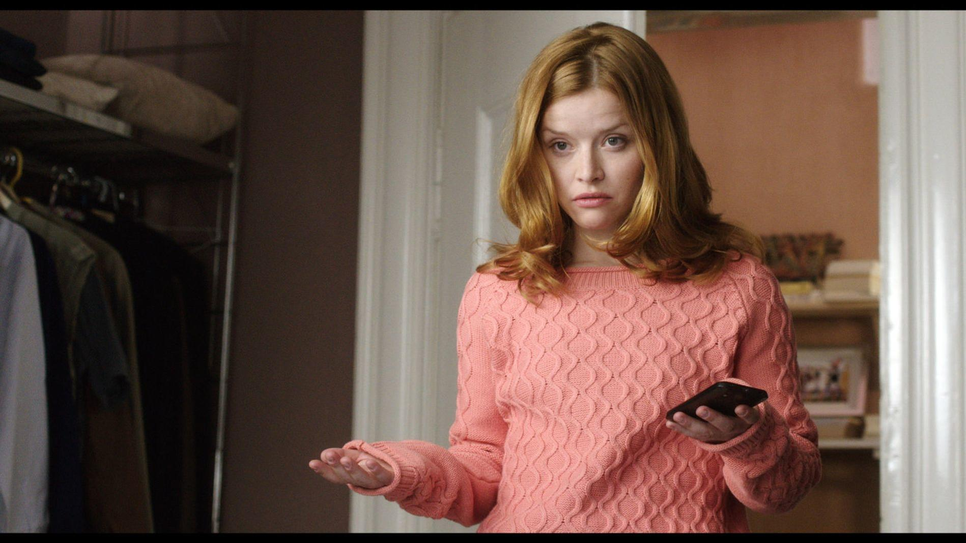 Eine Frau mit roten Haaren in einem rosafarbenen Pullover hält ein Handy.