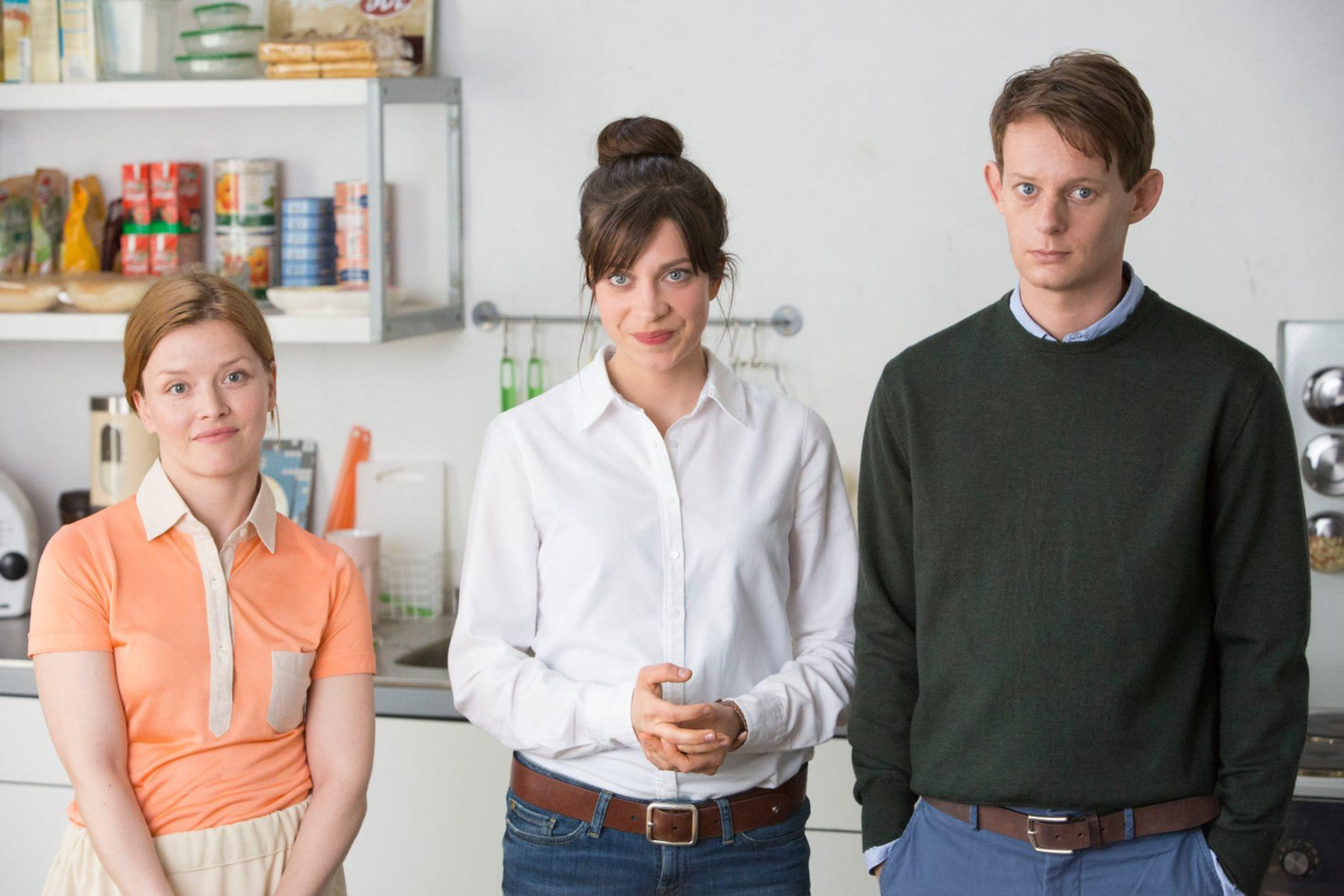 Drei Personen stehen in einer Küche. Links eine Frau in Orange, Mitte eine Frau in Weiß, rechts ein Mann in Grün.