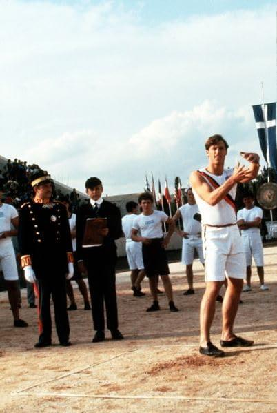 Ein Mann in weißer Sportkleidung hält eine Scheibe, umgeben von anderen Athleten und Würdenträgern.
