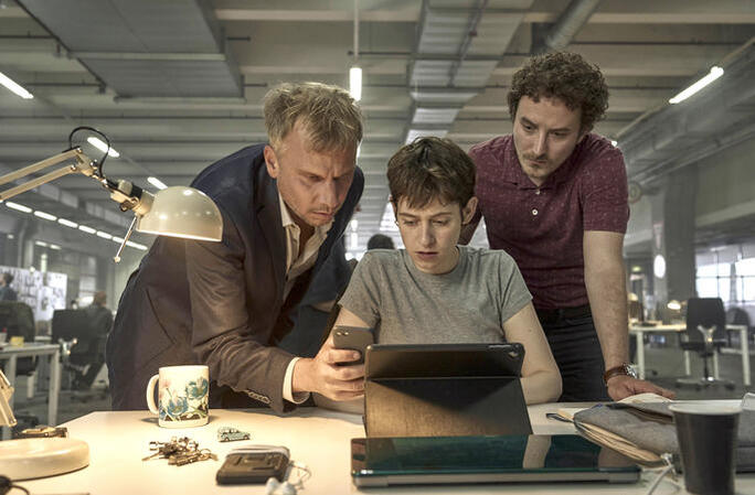 Drei Personen blicken auf einen Tablet-Bildschirm, zwei stehen, einer sitzt. Büro im Hintergrund.