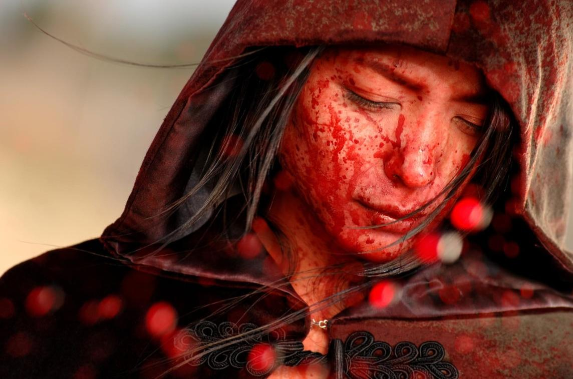 Eine Person mit blutverschmiertem Gesicht unter einer Kapuze. Unscharfe rote Lichter im Vordergrund.