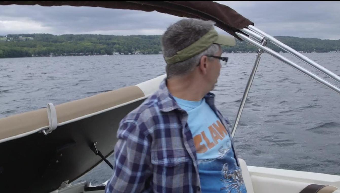 Ein Mann auf einem Boot blickt über das Wasser auf eine bewaldete Küste.