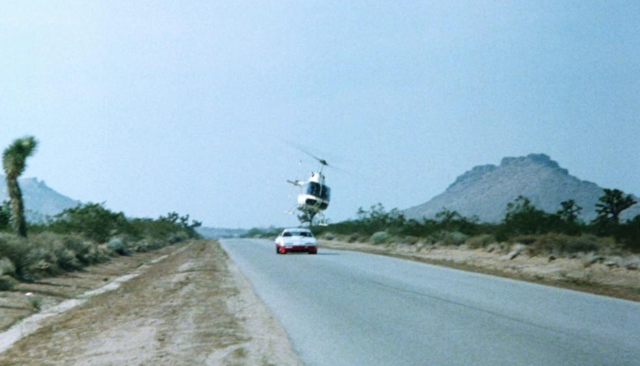 Ein weißer Helikopter fliegt über einem weißen und roten Auto auf einer Wüstenstraße.