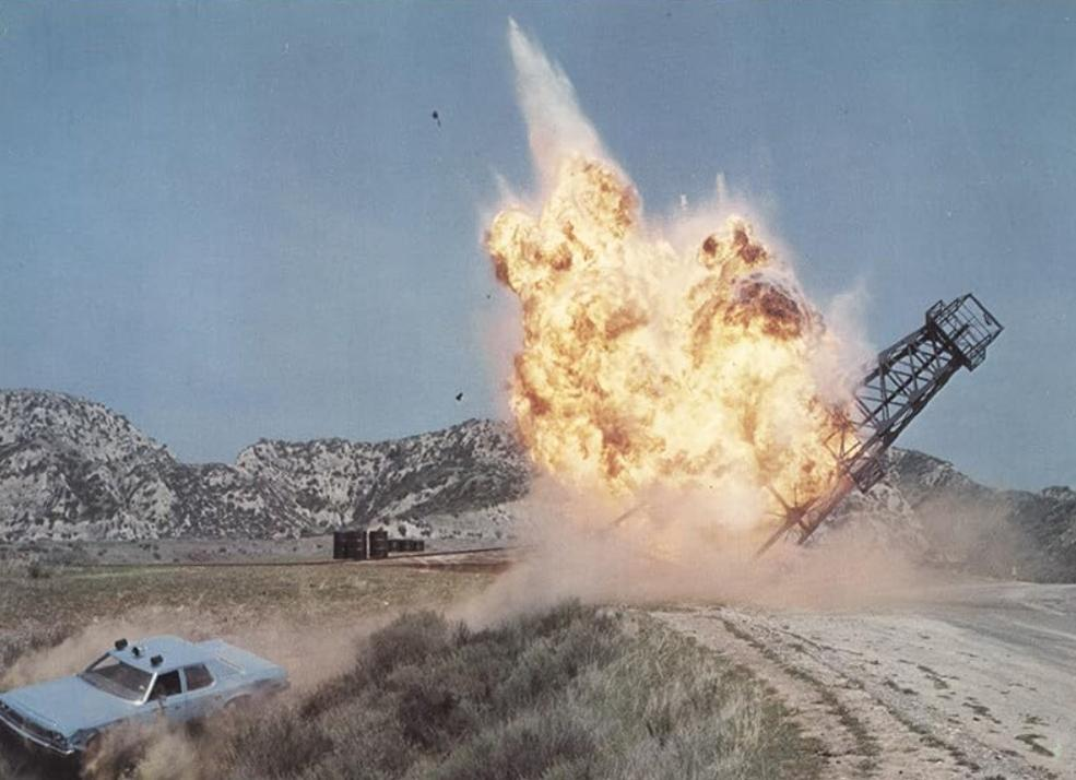 Ein Auto rast auf einer staubigen Straße, während im Hintergrund eine massive Explosion einen Turm zerstört.