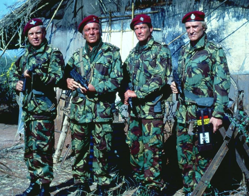 Vier Soldaten in Tarnanzügen und roten Baretts stehen nebeneinander und halten Waffen.