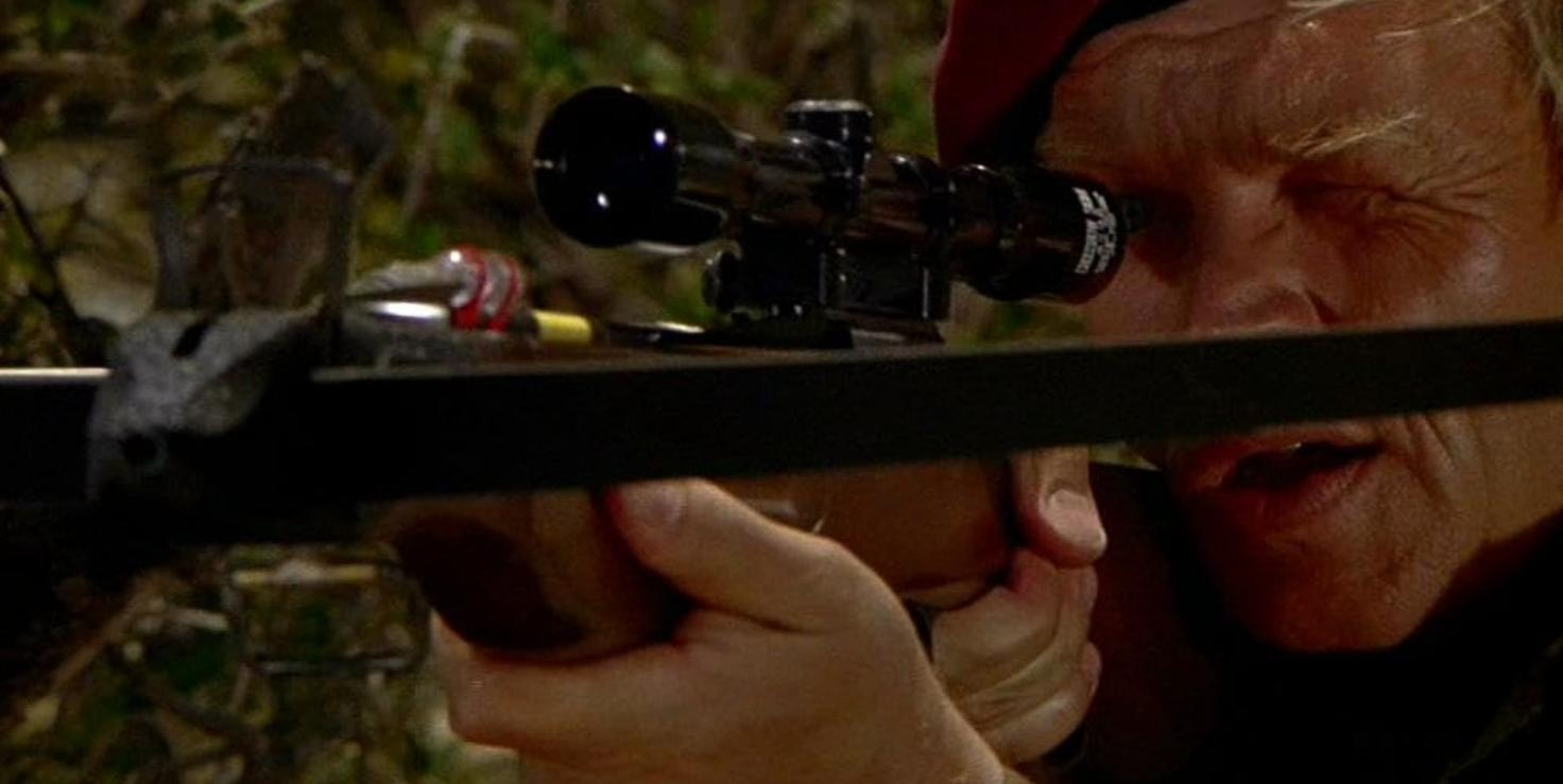Ein Mann mit hellem Haar zielt mit einem Gewehr im Wald.