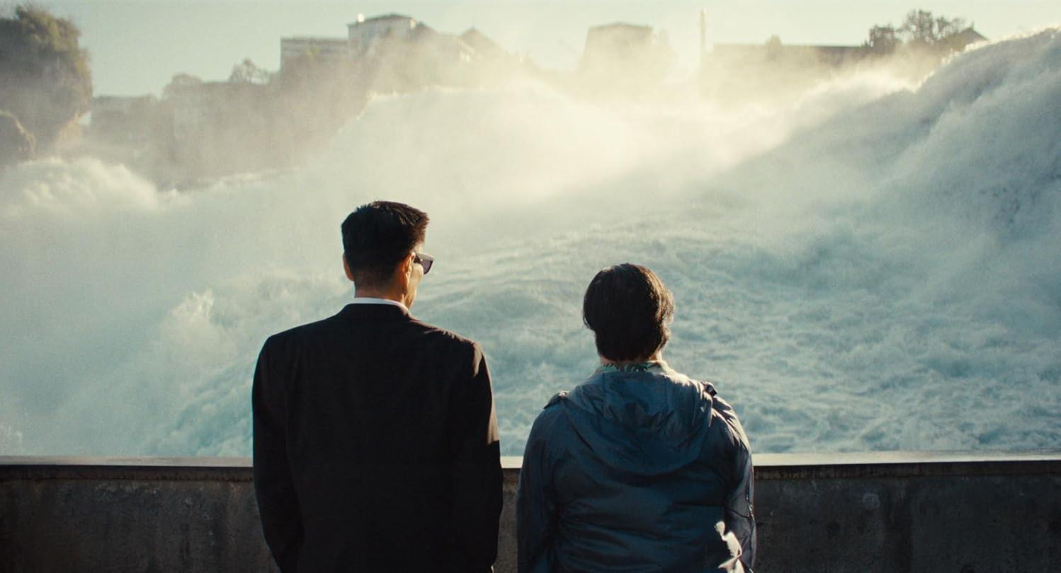 Zwei Personen stehen vor einem tosenden Wasserfall, Gebäude im Hintergrund.